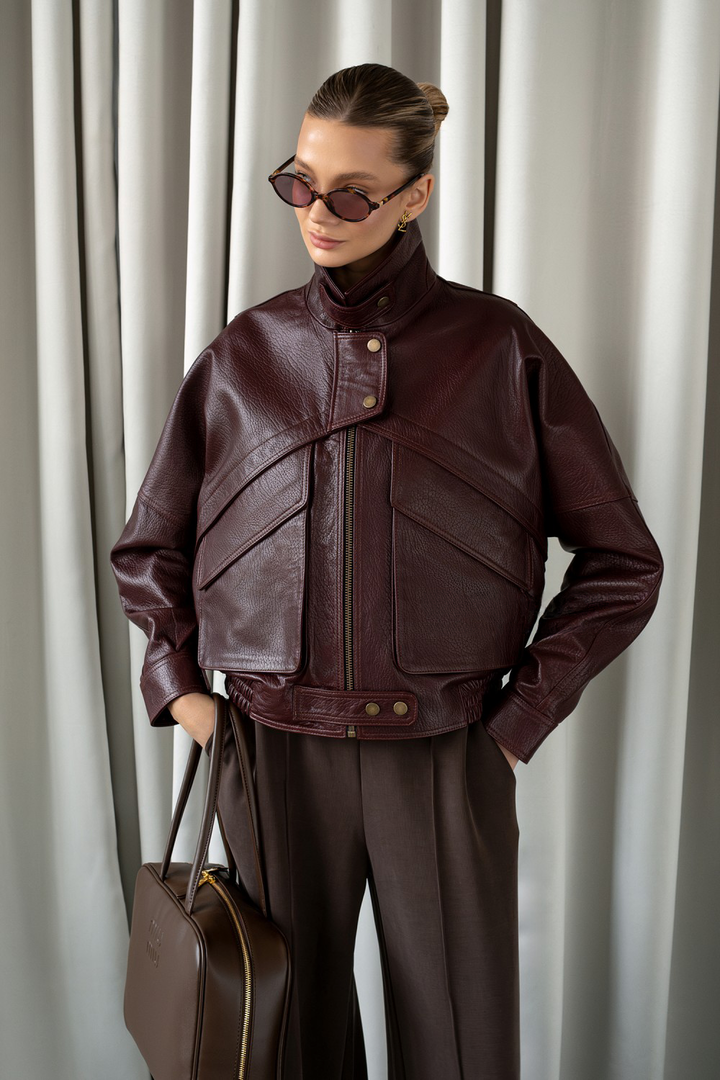 Woman in structured burgundy leather jacket styled with wide-leg trousers and sunglasses, showcasing bold luxury outerwear.