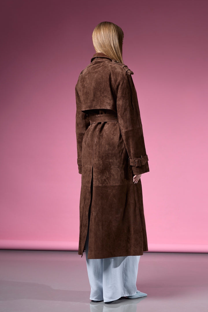 A woman models a chocolate brown trench coat in premium suede leather, designed with a figure-flattering shape and a sleek double-breasted closure.